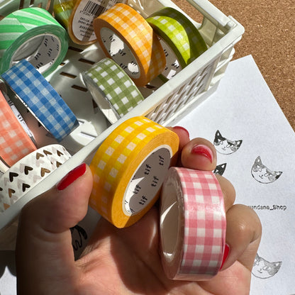 Assorted colorful washi tape rolls in a basket with a hand holding a roll of pink checkered tape.