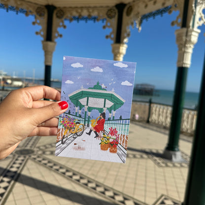 Hand holding a postcard with a scenic view of a pavilion and sea in the background