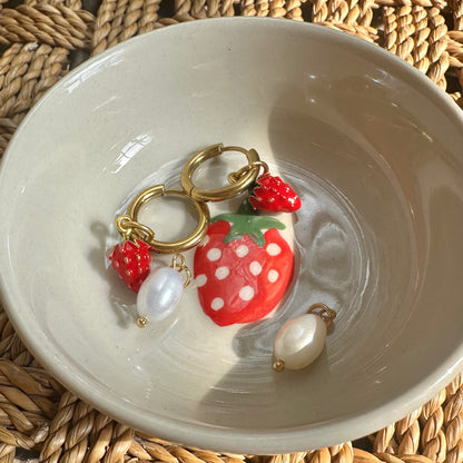Strawberry-themed earrings with gold accents in a white bowl on a woven surface