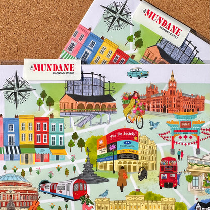 Two printed art cards or posters featuring colorful illustrated London landmarks and buildings, laid on a cork background. The cards show scenes such as a red double-decker bus, tube station, cathedral, market stall, and compass rose, with the title 'The MUNDANE BY LONDYN STUDIO' visible on each card.