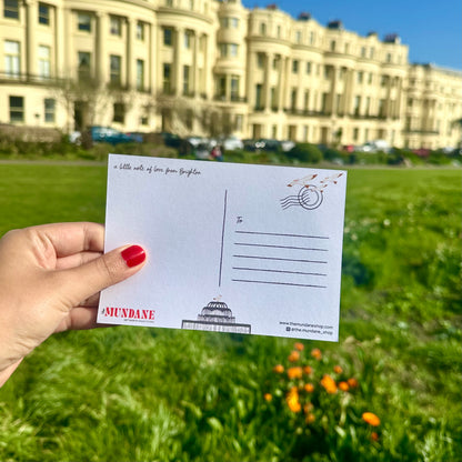 Hand holding a postcard with 'Mundane' branding in a park setting