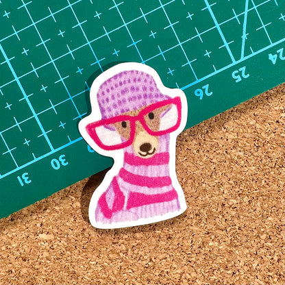 Sticker of a bear wearing a pink hat and scarf on a cork surface with a green cutting mat in the background.