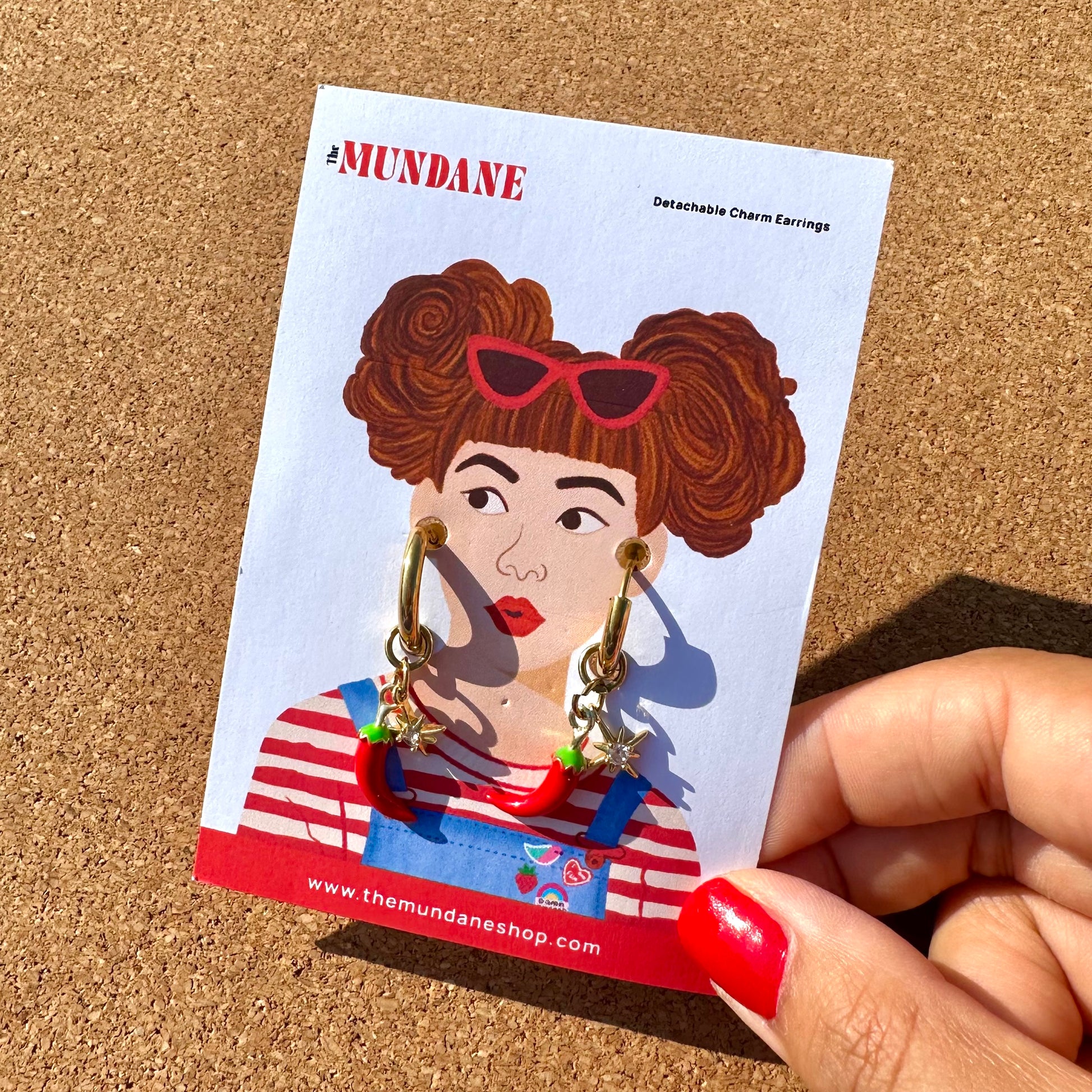 A hand with red nail polish holds a card displaying gold charm earrings. The card features a retro-style illustration of a woman with curly brown hair, red sunglasses, and a red-and-white striped shirt.