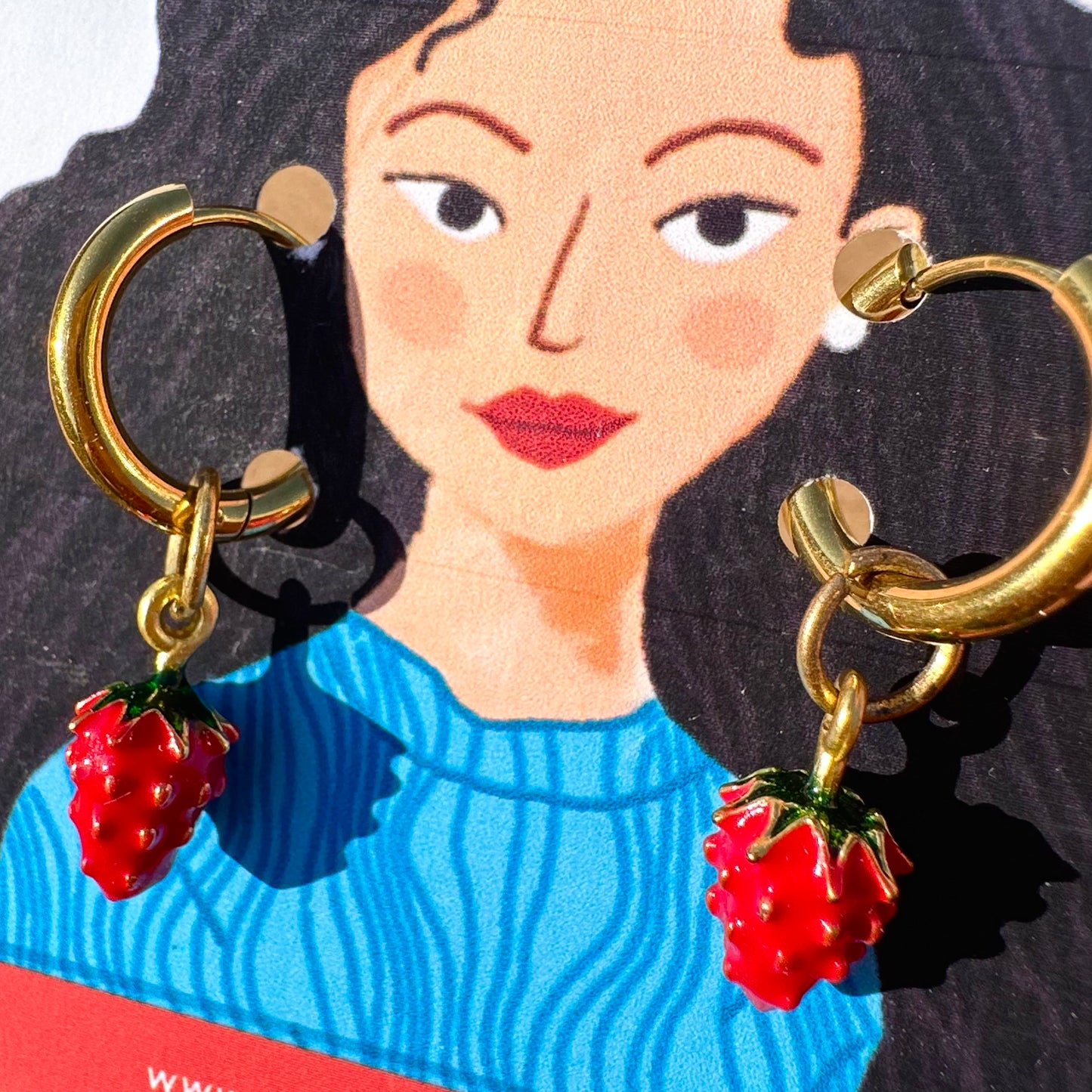 Gold hoop earrings with dangling red strawberry charms, shown against a stylized illustrated card featuring a woman in a blue sweater.