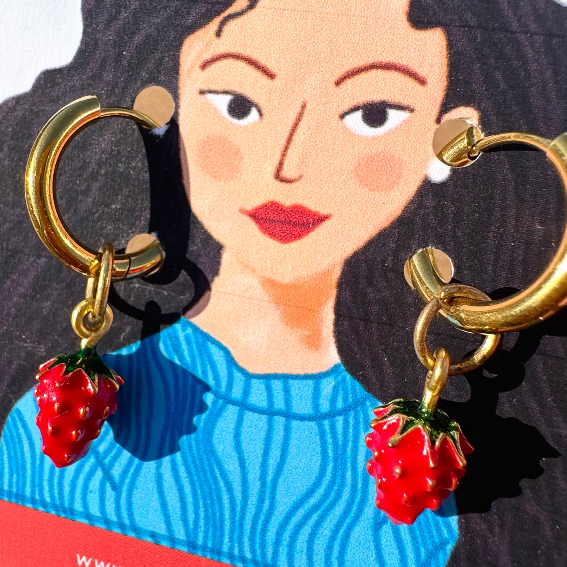 Gold hoop earrings with dangling red strawberry charms, shown against a stylized illustrated card featuring a woman in a blue sweater.