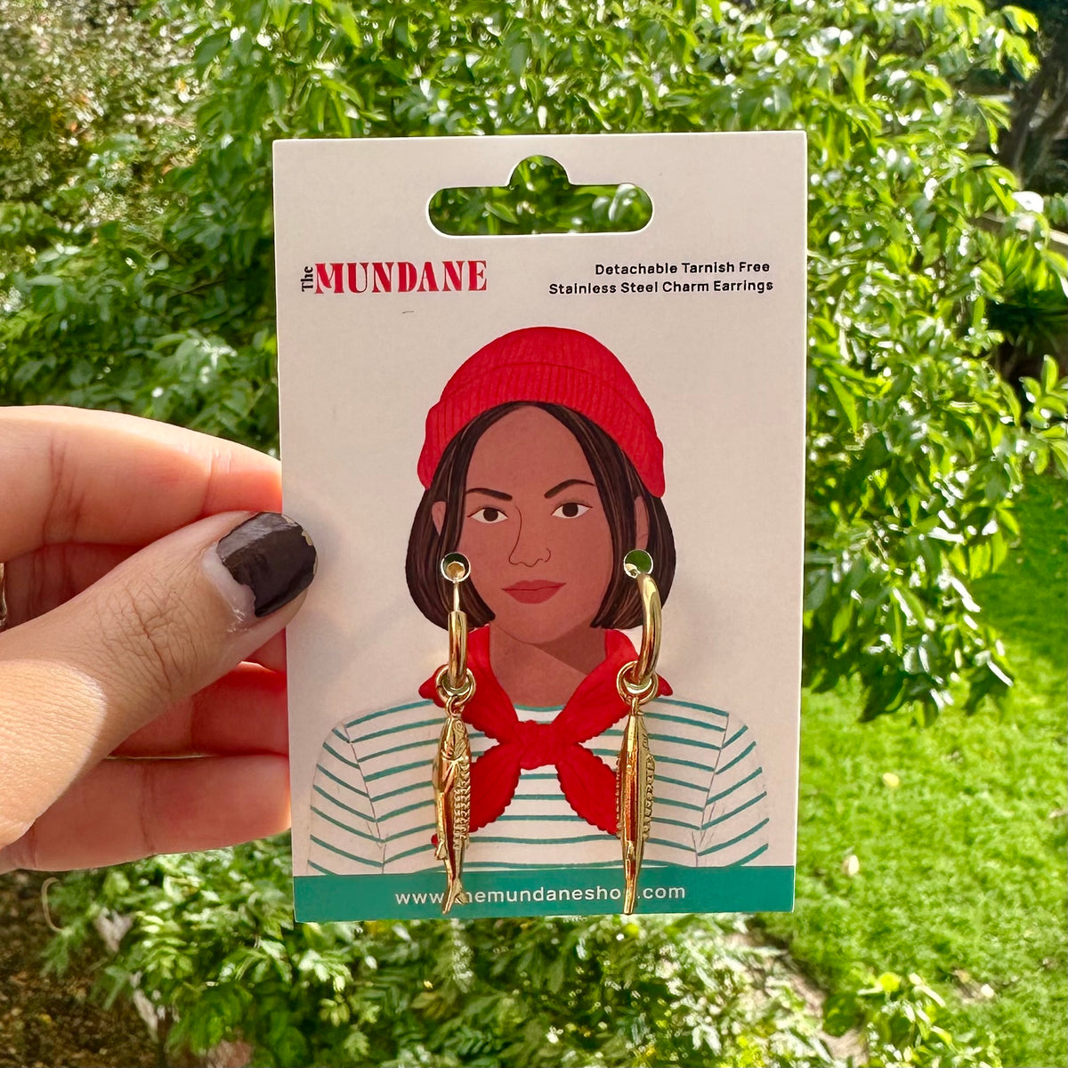A hand holds a card displaying gold-toned dangling earrings with red and green details. The card is printed with an illustration of a woman wearing a red beanie and red neck scarf, and the brand name MUNDANE is visible at the top.