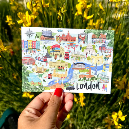 A hand with red nail polish holds a square illustrated London postcard or art print in front of a blurred background of yellow flowers. The postcard features colorful drawings of London landmarks, buses, bridges, and buildings, with the word 'London' printed at the bottom.