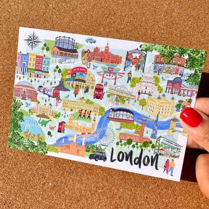 A hand with red nail polish holds a white illustrated postcard featuring colorful London landmarks and scenes, with the word 'London' written at the bottom, against a brown cork background.