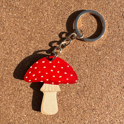 A mushroom-shaped keychain with a red cap covered in white spots, a light natural wood stem, and a silver metal key ring, placed on a brown cork background.