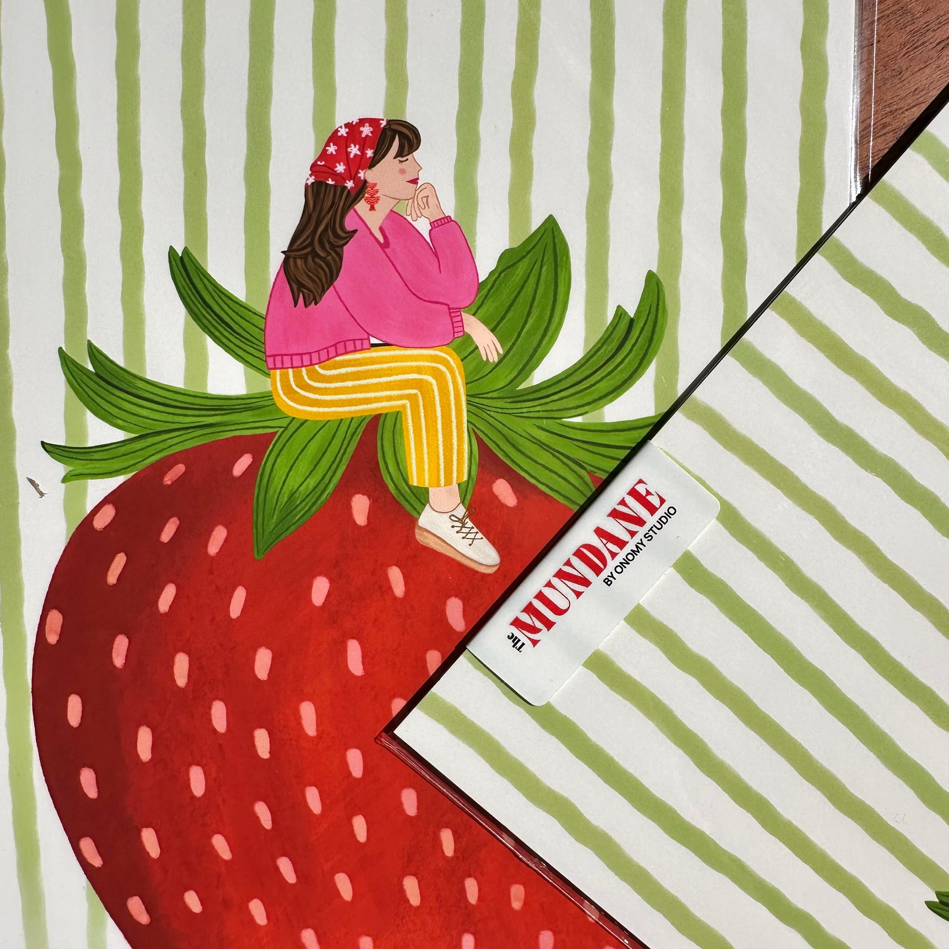 Illustration of a person sitting on a strawberry with green leaves, against a striped background. The brand 'Mundane' is visible.