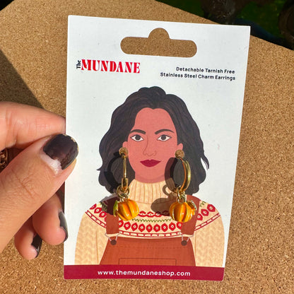 A hand holds a card displaying a pair of dangling earrings. The card shows the brand name 'The Mundane' and an illustration of a woman wearing patterned overalls.