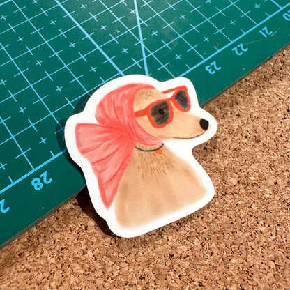 Sticker of a dog with a pink bow and sunglasses on a cork surface with a ruler in the background.