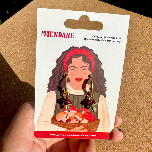 A hand holding a white display card featuring an illustration of a woman wearing red charm earrings. The card reads 'The Mundane' and 'Detachable Tarnish Free Stainless Steel Charm Earrings,' with the earrings pictured dangling below the illustrated figure.