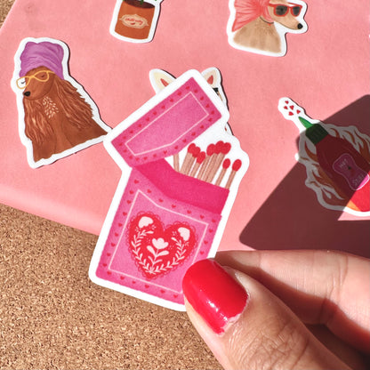 Sticker of a pink matches box with a heart design on a pink background.