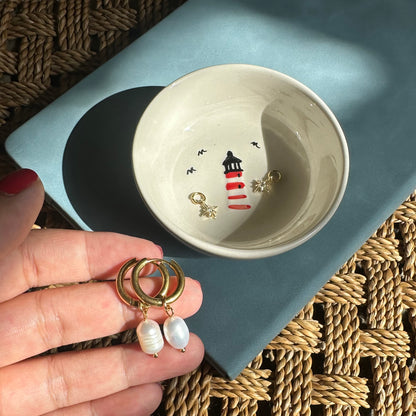 Hand holding gold hoop earrings with pearls near a ceramic bowl with a lighthouse design on a textured surface.