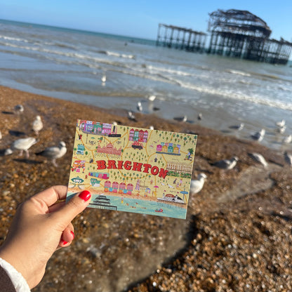 Hand holding a postcard with 'Brighton' on it against a beach background with seagulls and the remains of a pier.