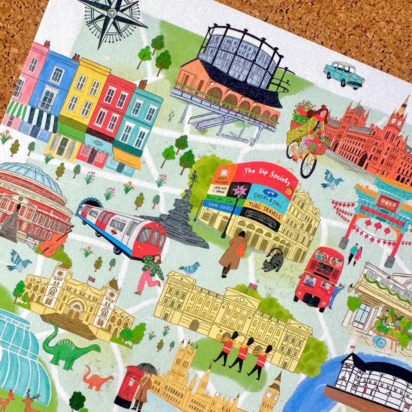A colorful illustrated map print featuring London landmarks and scenes, including The Shard, Big Ben, a red double-decker bus, a red train, a cyclist, and pastel-colored buildings, shown laid flat on a cork background.