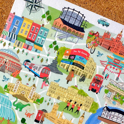 A colorful illustrated map print featuring London landmarks and scenes, including The Shard, Big Ben, a red double-decker bus, a red train, a cyclist, and pastel-colored buildings, shown laid flat on a cork background.