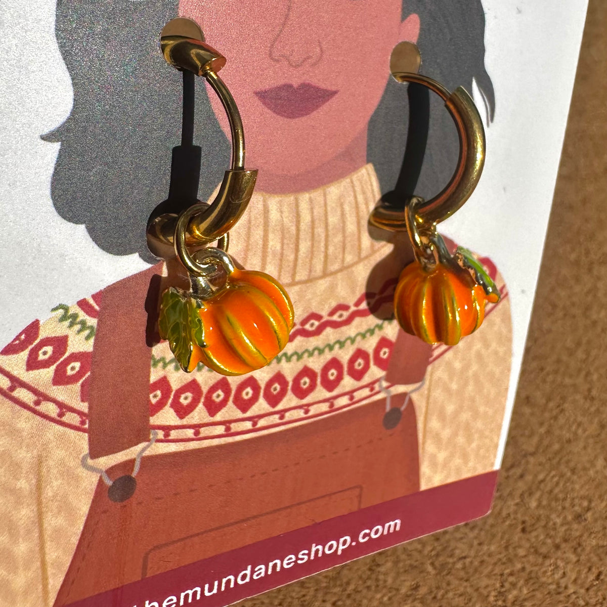 Gold-tone hoop earrings with small orange and yellow striped pumpkin charms and green leaves, displayed on a printed card.
