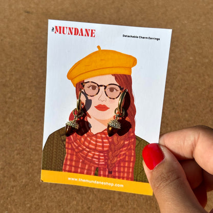 A hand with red nail polish holds a white card featuring an illustration of a woman wearing large gold and dark dangling earrings. The card is branded MUNDANE and reads 'Detachable Charm Earrings.