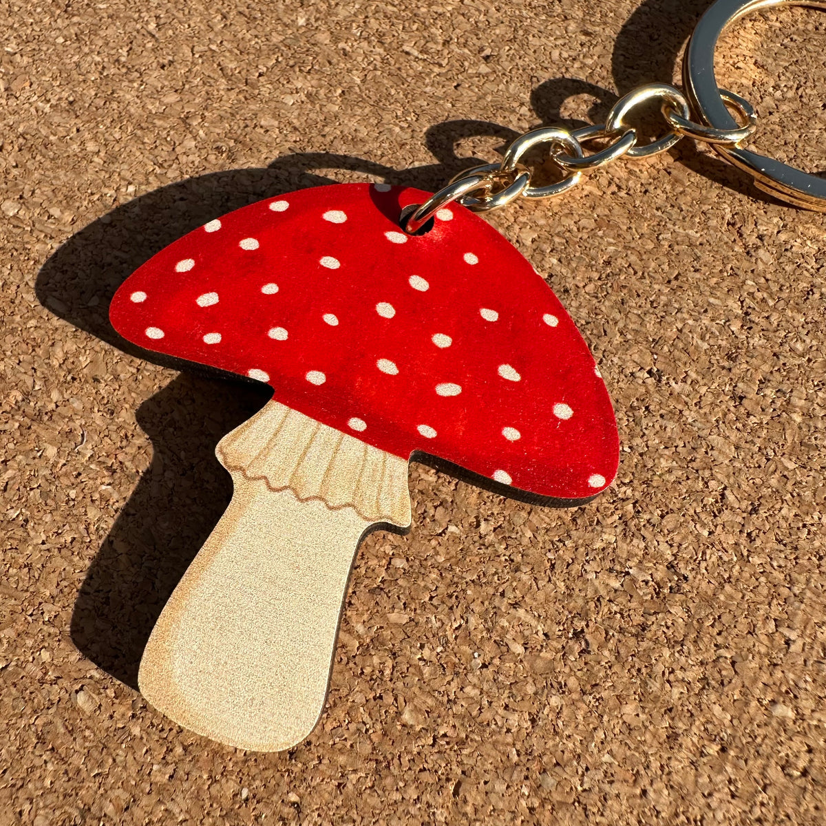 A mushroom-shaped keychain with a red cap, white polka dots, and a natural wood stem, attached to a silver metal key ring, resting on a cork surface.