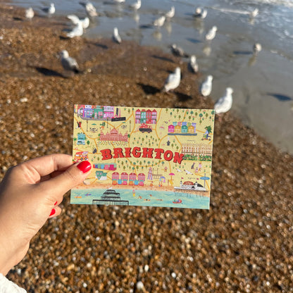 Hand holding a postcard with 'Brighton' illustration on a beach with seagulls.