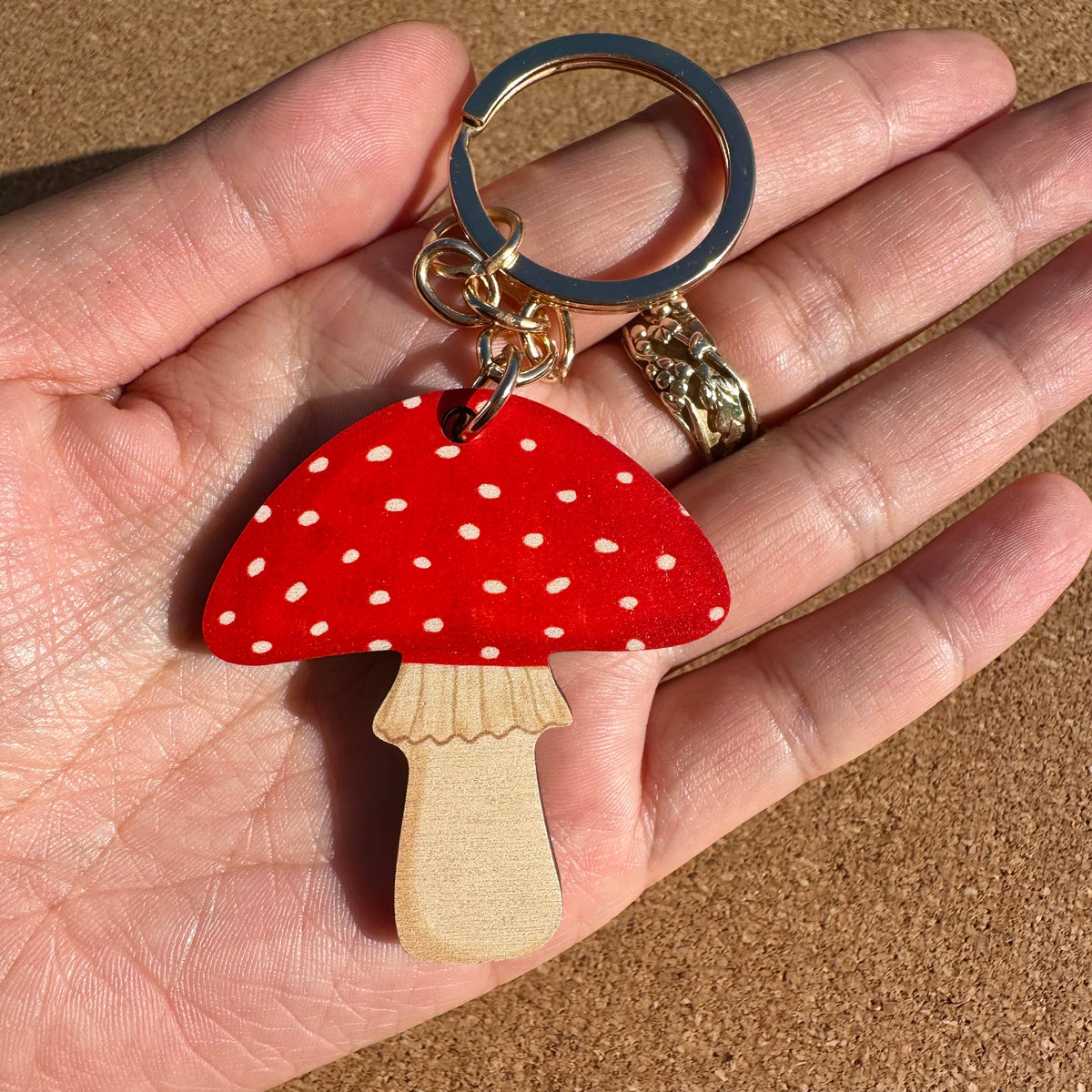 A red mushroom-shaped keychain with white polka dots and a natural wood stem, held in a person's hand. The keychain has a black split ring and gold-tone metal hardware.