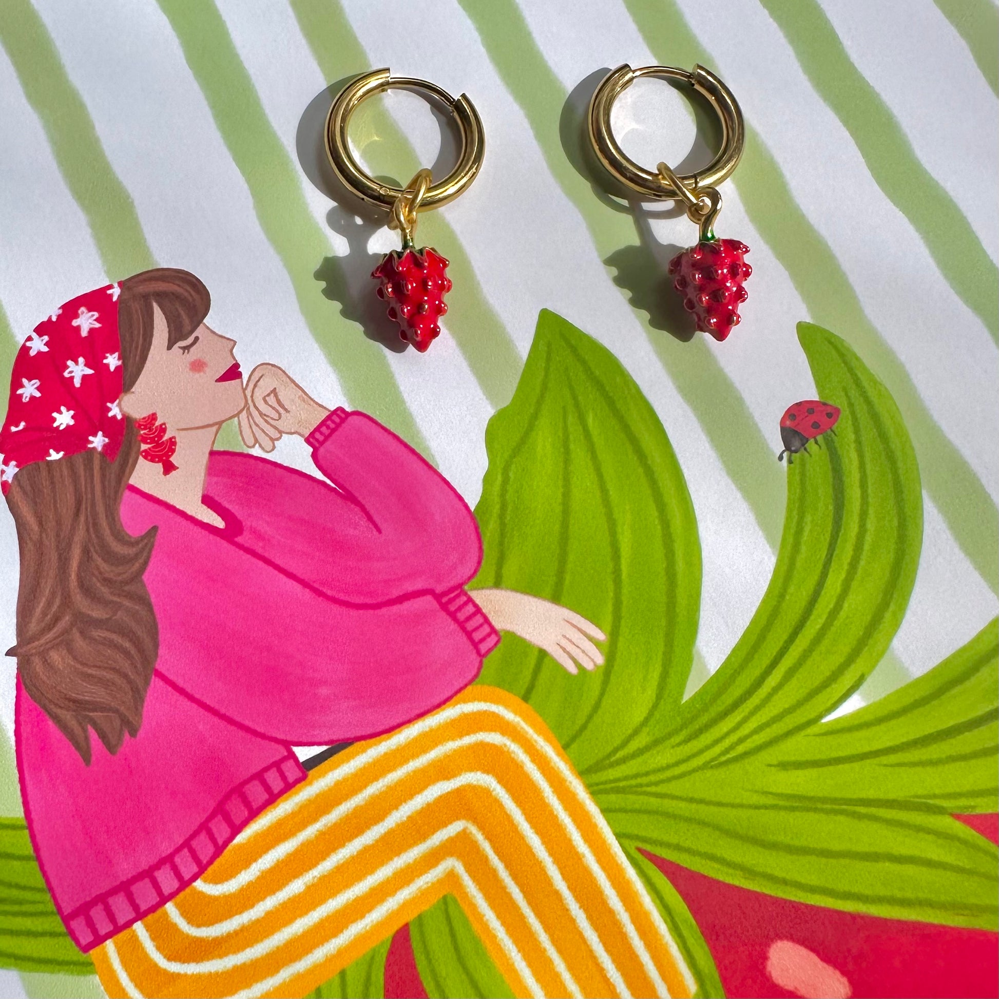 A pair of gold-tone hoop earrings with small red beaded strawberry charms, displayed on a colorful illustrated background featuring a woman in a pink sweater and yellow striped pants.