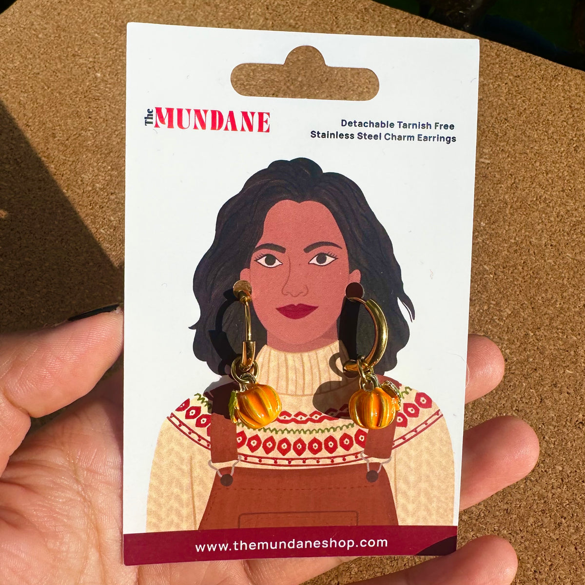 A hand holding a card backing for The Mundane detachable tarnish-free stainless steel charm earrings. The card features an illustration of a woman wearing orange pumpkin charms and gold hoop earrings, with the text 'The MUNDANE' and 'Detachable Tarnish Free Stainless Steel Charm Earrings' on a corkboard-like background.