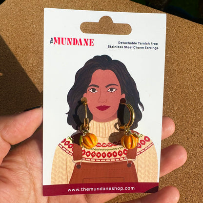 A hand holding a card backing for The Mundane detachable tarnish-free stainless steel charm earrings. The card features an illustration of a woman wearing orange pumpkin charms and gold hoop earrings, with the text 'The MUNDANE' and 'Detachable Tarnish Free Stainless Steel Charm Earrings' on a corkboard-like background.