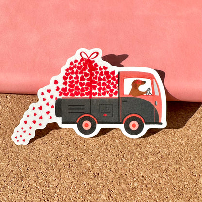 Sticker of a truck carrying hearts on a cork surface with a pink background