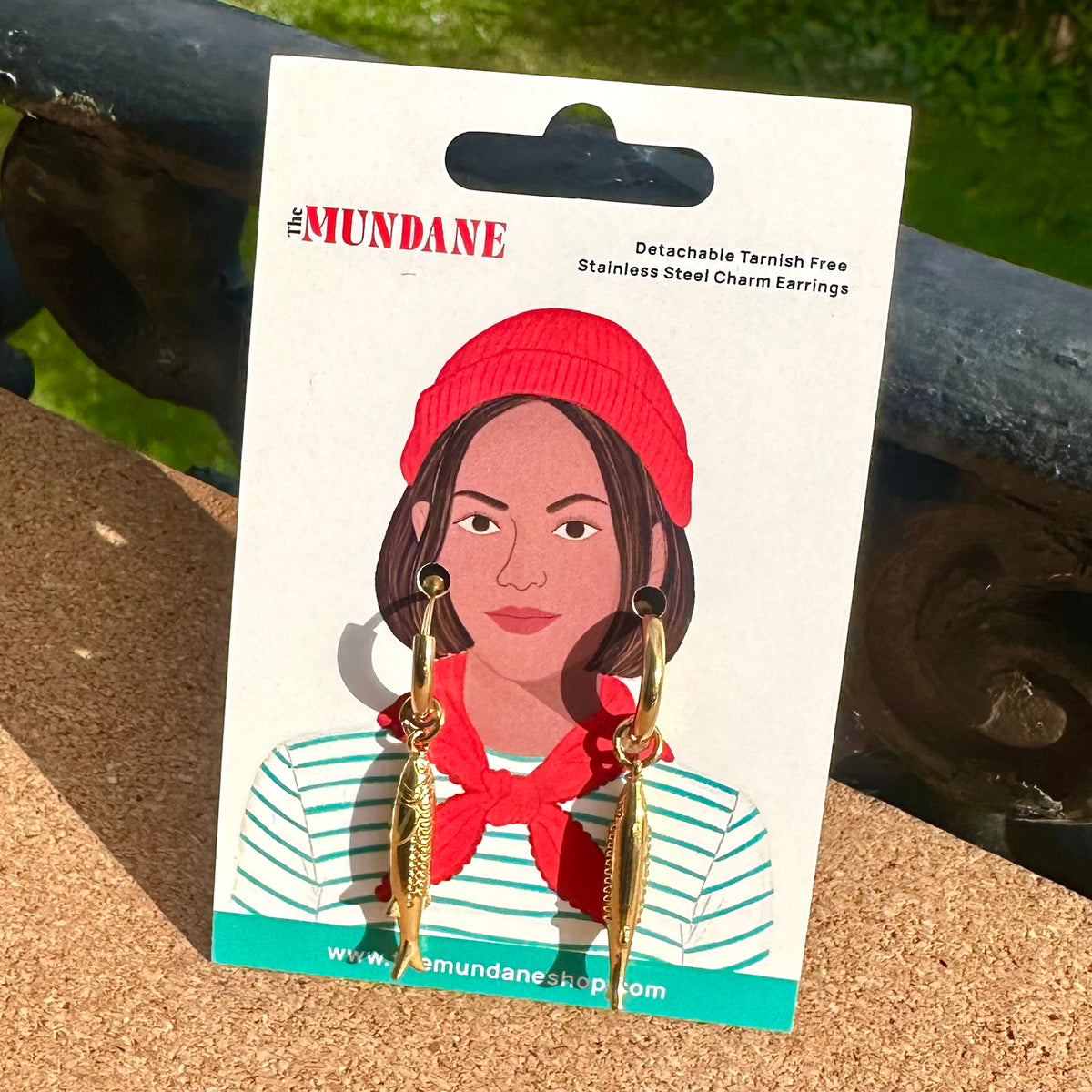 The Mundane brand stainless steel charm earrings displayed on a white illustrated backing card. The earrings feature gold-toned fish charms with dark metal tops and black eye details.
