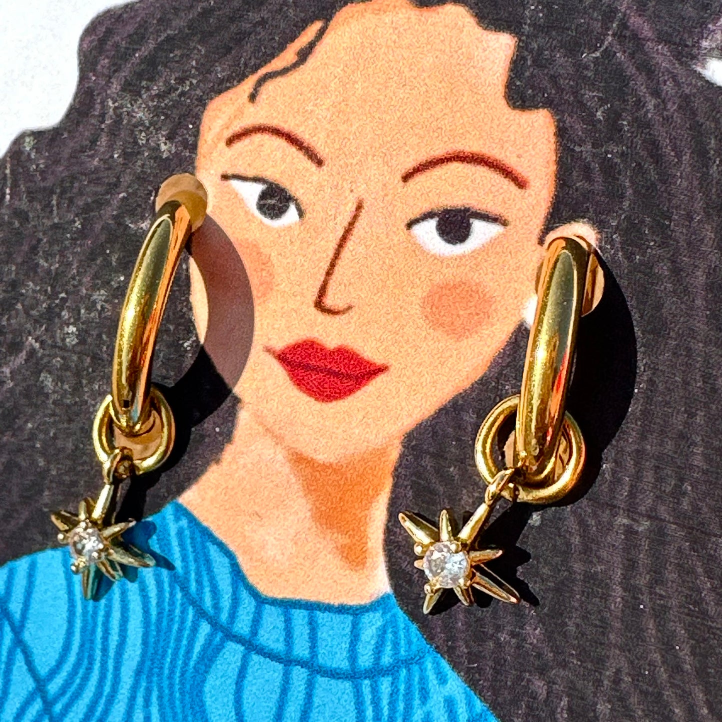 A close-up of a person wearing a pair of gold hoop earrings with dangling starburst charms. The image is cropped to show the person's face, hair, and upper torso, with the earrings clearly visible against dark hair and a blue top.