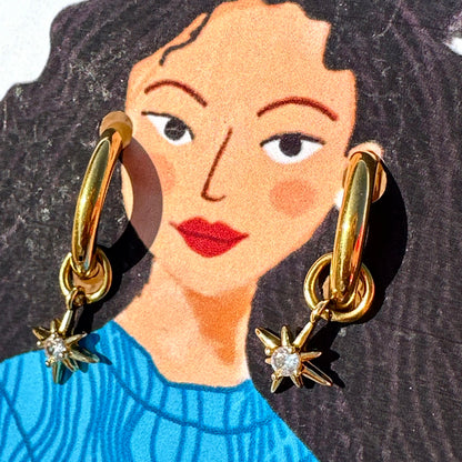 A close-up of a person wearing a pair of gold hoop earrings with dangling starburst charms. The image is cropped to show the person's face, hair, and upper torso, with the earrings clearly visible against dark hair and a blue top.