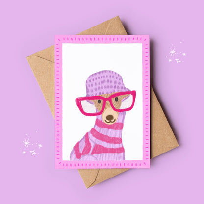 Greeting card with a llama wearing pink glasses and a sweater, next to a brown envelope on a purple background.