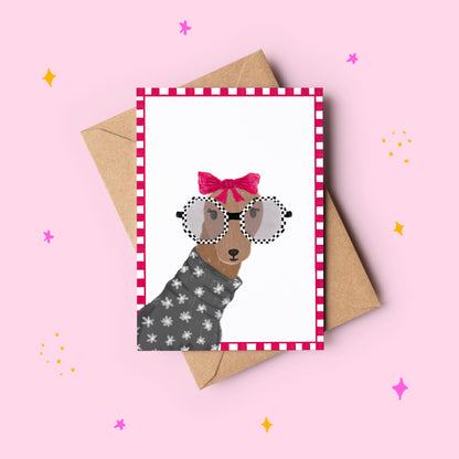 Greeting card with a dog illustration wearing sunglasses and a bow, next to a brown envelope on a pink background.