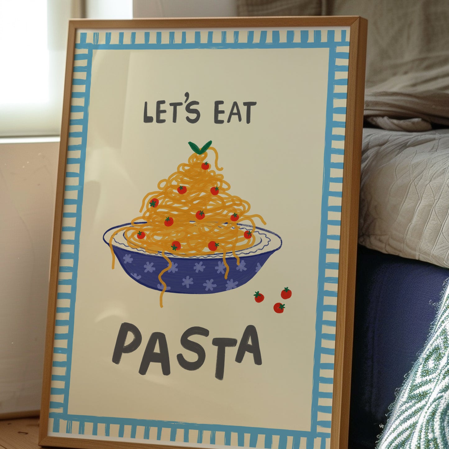 A framed art print featuring a hand-drawn illustration of spaghetti in a blue bowl with the text 'LET'S EAT PASTA' in dark gray lettering. The print has a cream background, a light blue patterned border, and is housed in a light brown wooden frame, leaning against a bed in a bedroom setting.