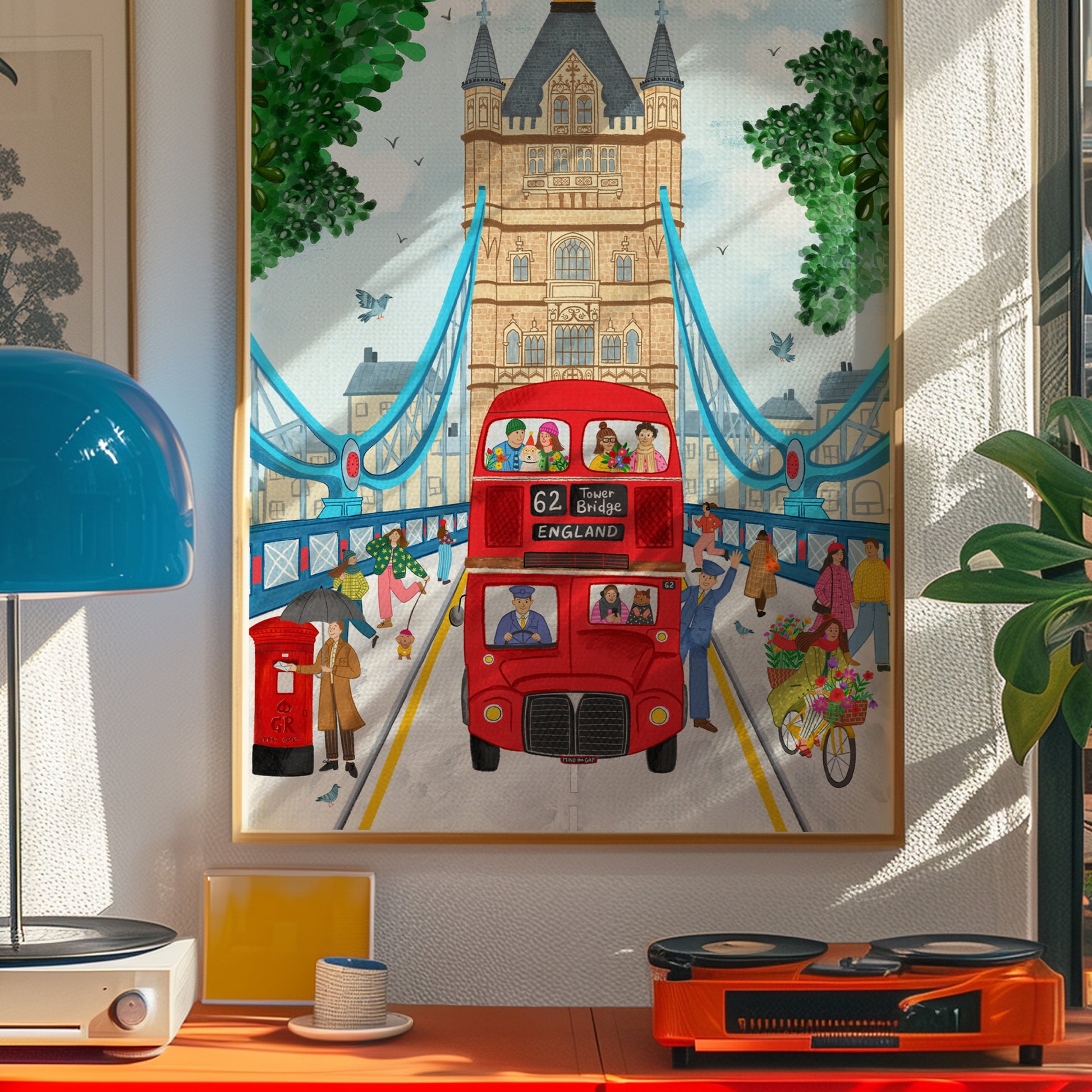A framed colorful art print depicting a London scene with Tower Bridge, a red double-decker bus, and pedestrians, displayed on a wall above a desk in a styled interior.