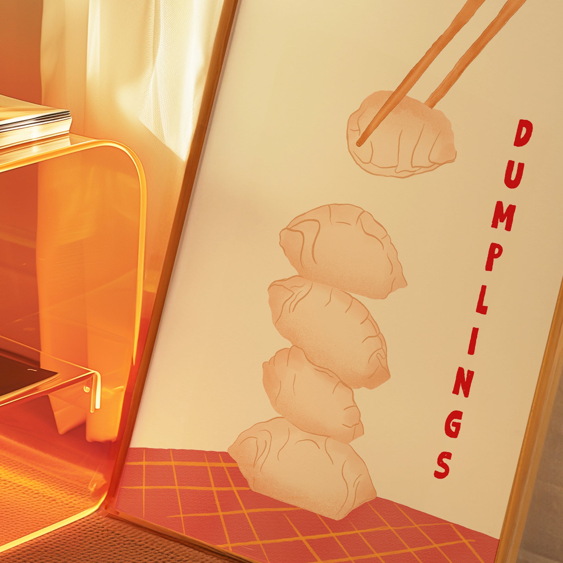 A framed art print featuring a minimalist illustration of four dumplings stacked vertically, with chopsticks lifting the top dumpling and the word 'DUMPLINGS' written vertically in red. The print is shown in a warm, orange-toned setting next to a modern glass shelf.