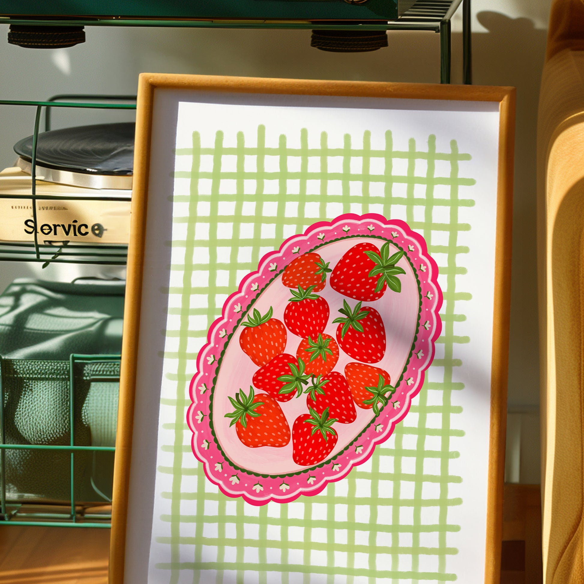 A framed art print featuring a whimsical illustration of red strawberries on a pink scalloped oval plate against a light green grid background, displayed in a warm indoor setting.