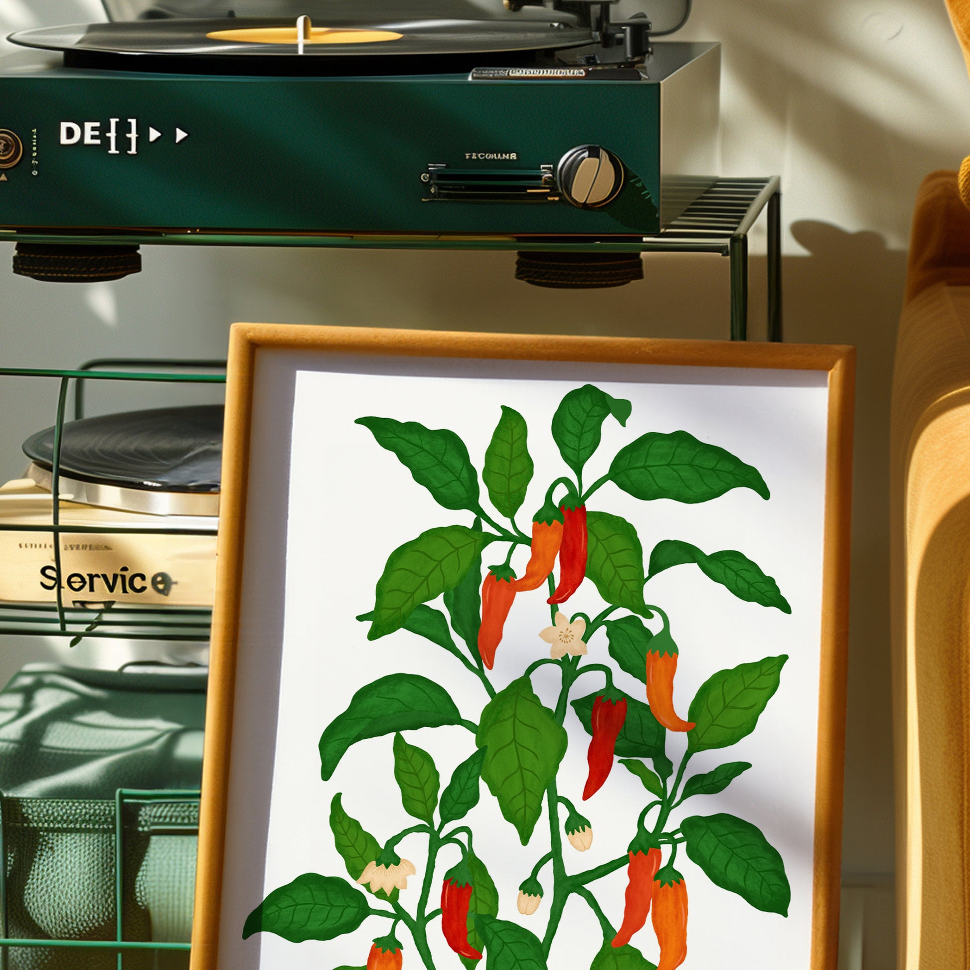 A framed art print featuring a stylized illustration of a chili pepper plant with green leaves and red-orange peppers on a white background, styled in a warm, sunlit room next to a green turntable.