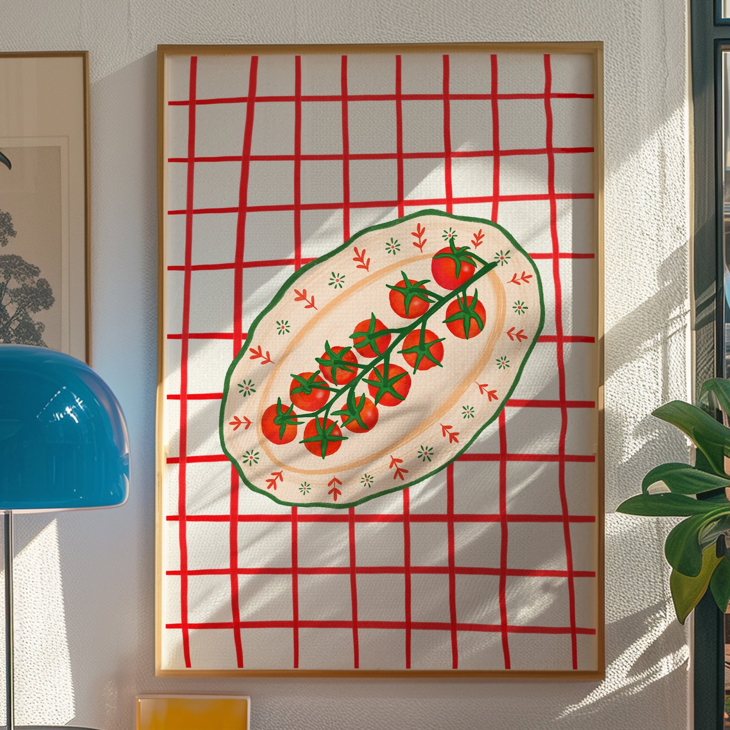 Framed wall art hanging on a textured white wall, featuring a red grid background with an oval plate of red tomatoes on a vine in the center. The frame is light wood, and surrounding decor includes a blue lamp on the left and a green plant on the right.