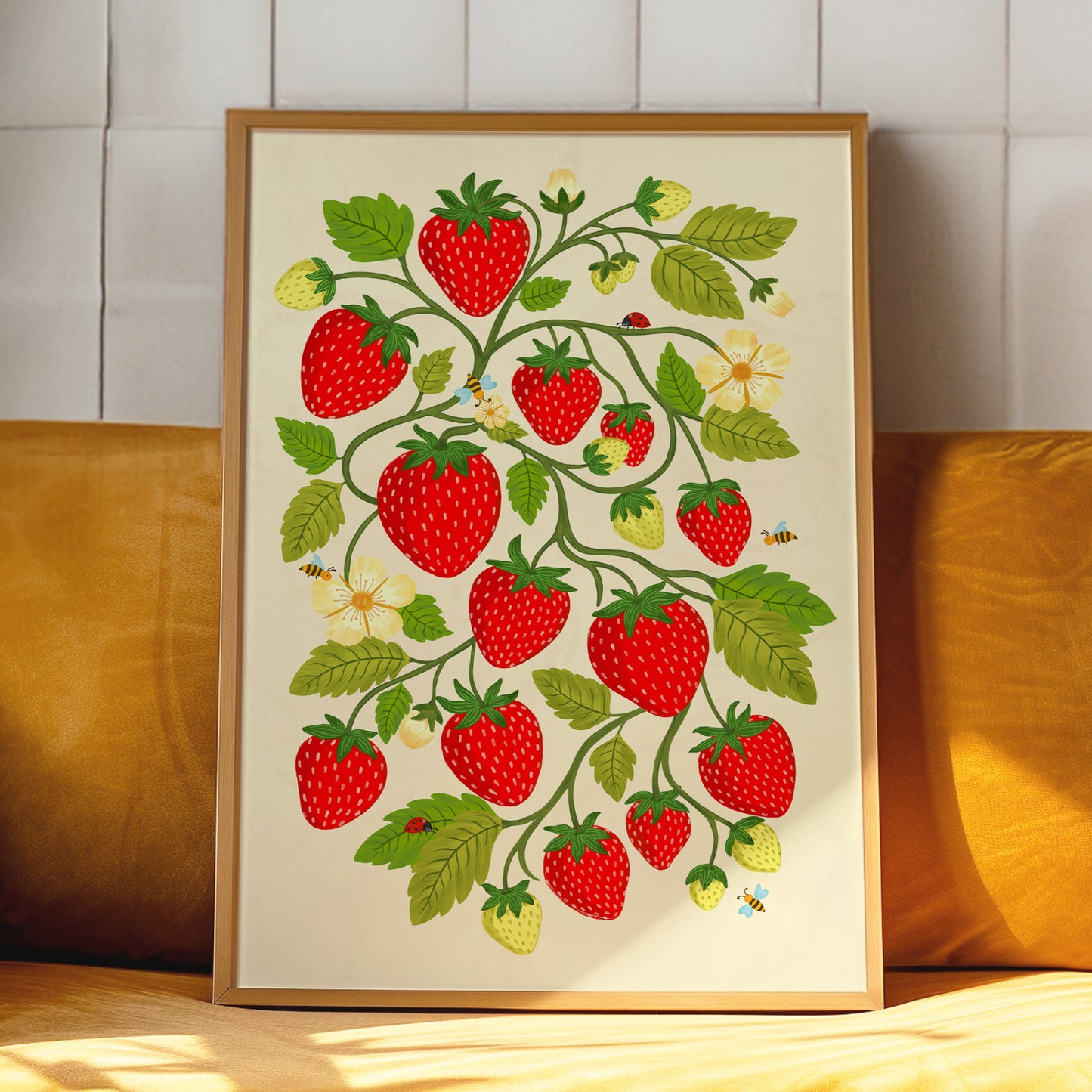 A framed art print featuring a whimsical illustration of red strawberries, green leaves, and small yellow flowers on a cream background, leaning against a yellow cushion in front of a white tiled wall.