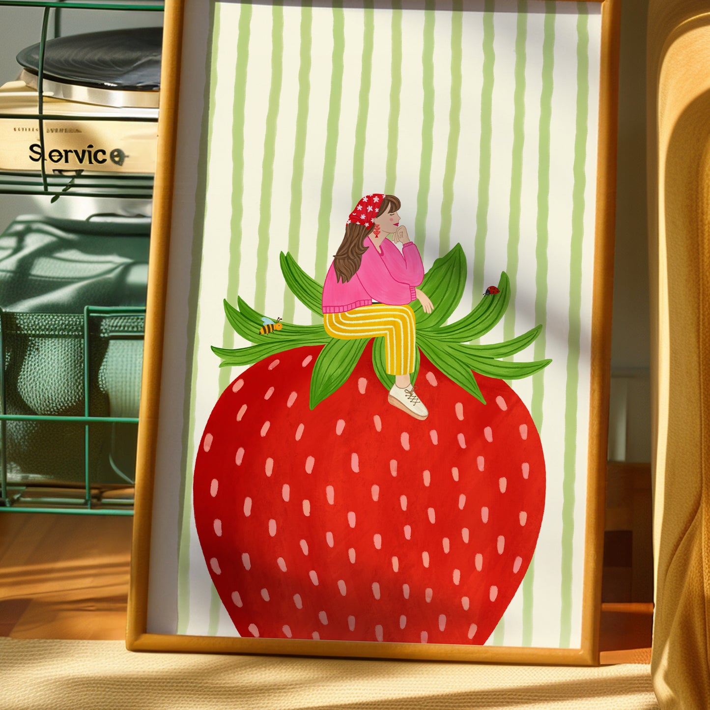 A framed art print featuring a whimsical illustration of a girl in a pink outfit sitting on a large red strawberry, set against a green and white striped background. The framed print is displayed on a wooden surface next to a green wire shelf.