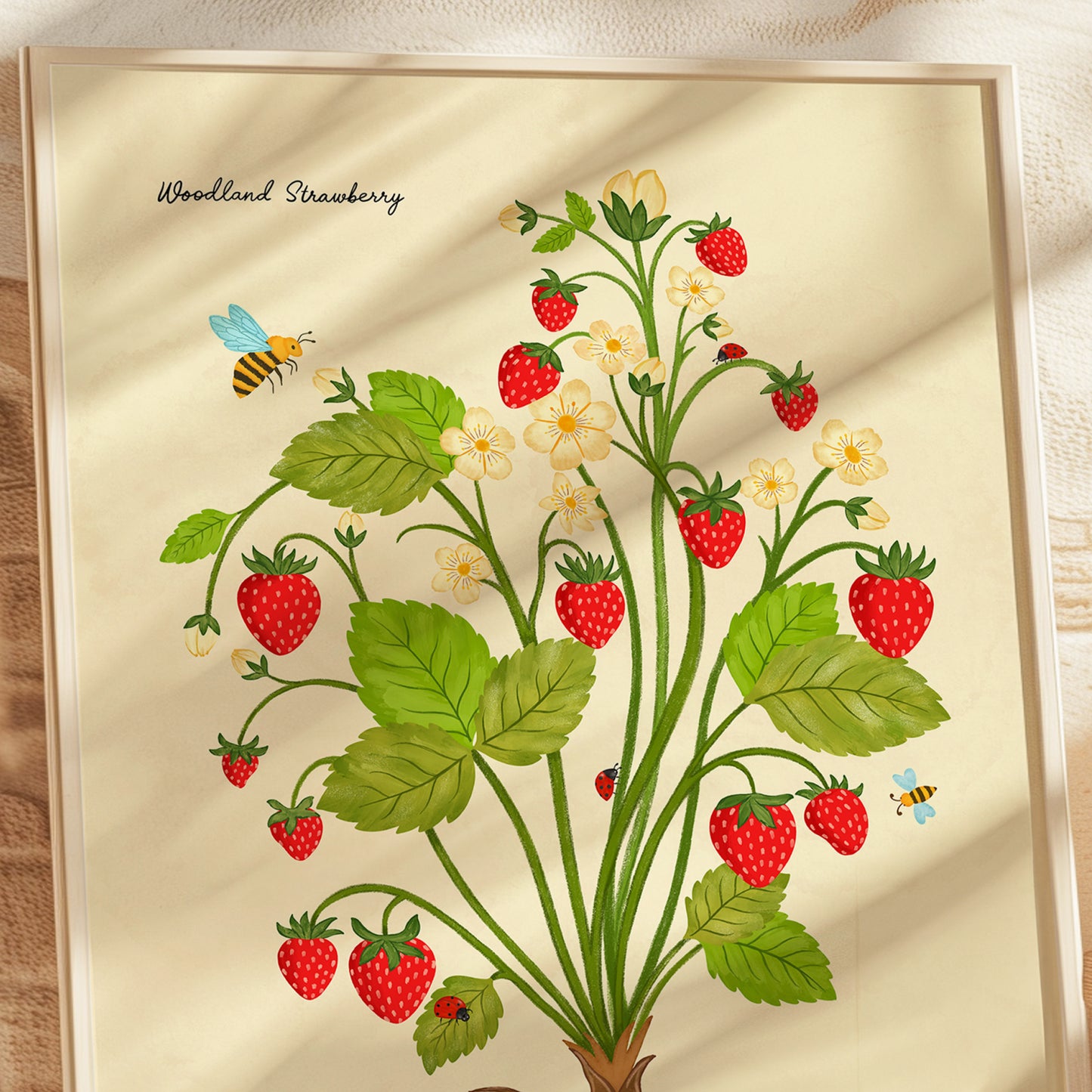 A framed botanical art print titled 'Woodland Strawberry' featuring red strawberries, green leaves, and small white flowers with bees on a cream background, shown in a light wood frame.