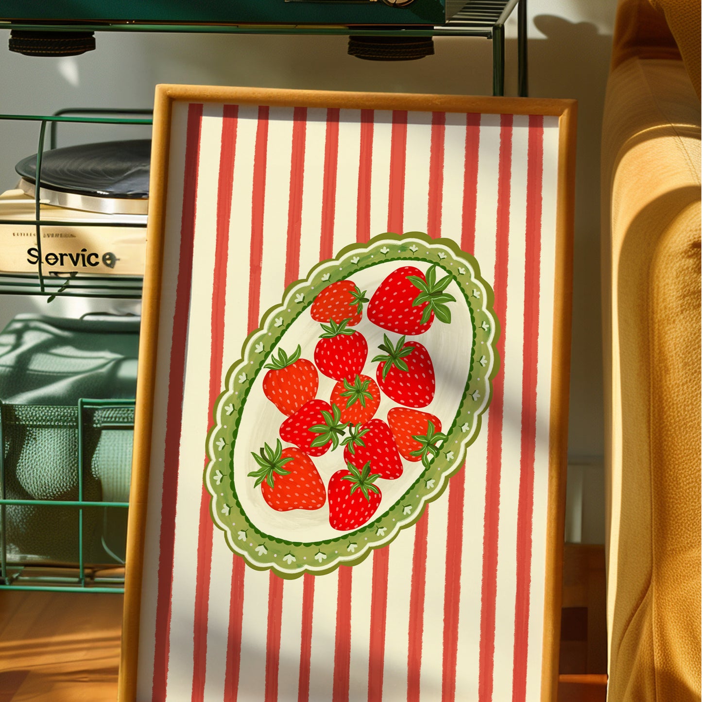 A framed art print featuring a vintage-style illustration of red strawberries on a white plate with a green scalloped border, set against a background of red and white vertical stripes. The frame is displayed in a warm, sunlit home environment.