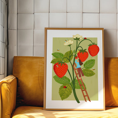 A framed art print featuring a whimsical illustration of a person climbing a ladder to harvest red strawberries from a plant with green leaves and white flowers, displayed on a yellow sofa against a white paneled wall.