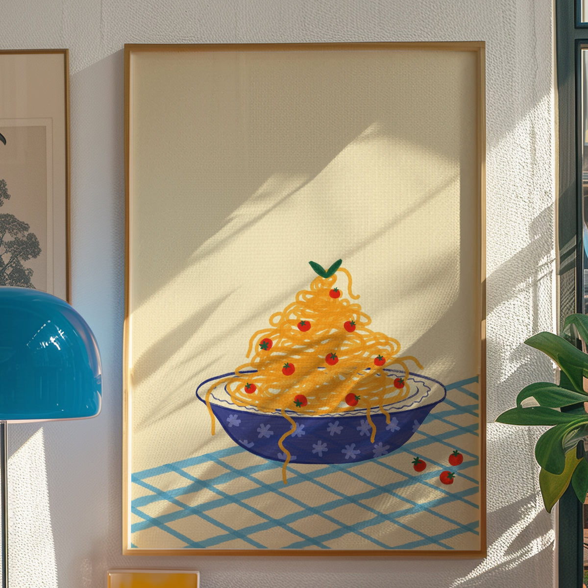 A framed art print depicting a whimsical bowl of yellow spaghetti with red cherry accents on a blue patterned surface, displayed on a textured white wall next to a blue lamp and green plant.