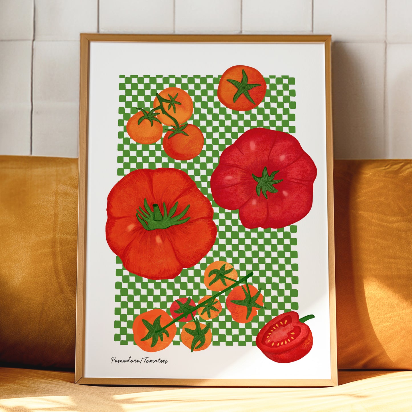 A framed art print featuring a colorful illustration of red and orange tomatoes on a green checkered background, leaning against a yellow cushion in front of a white tiled wall.