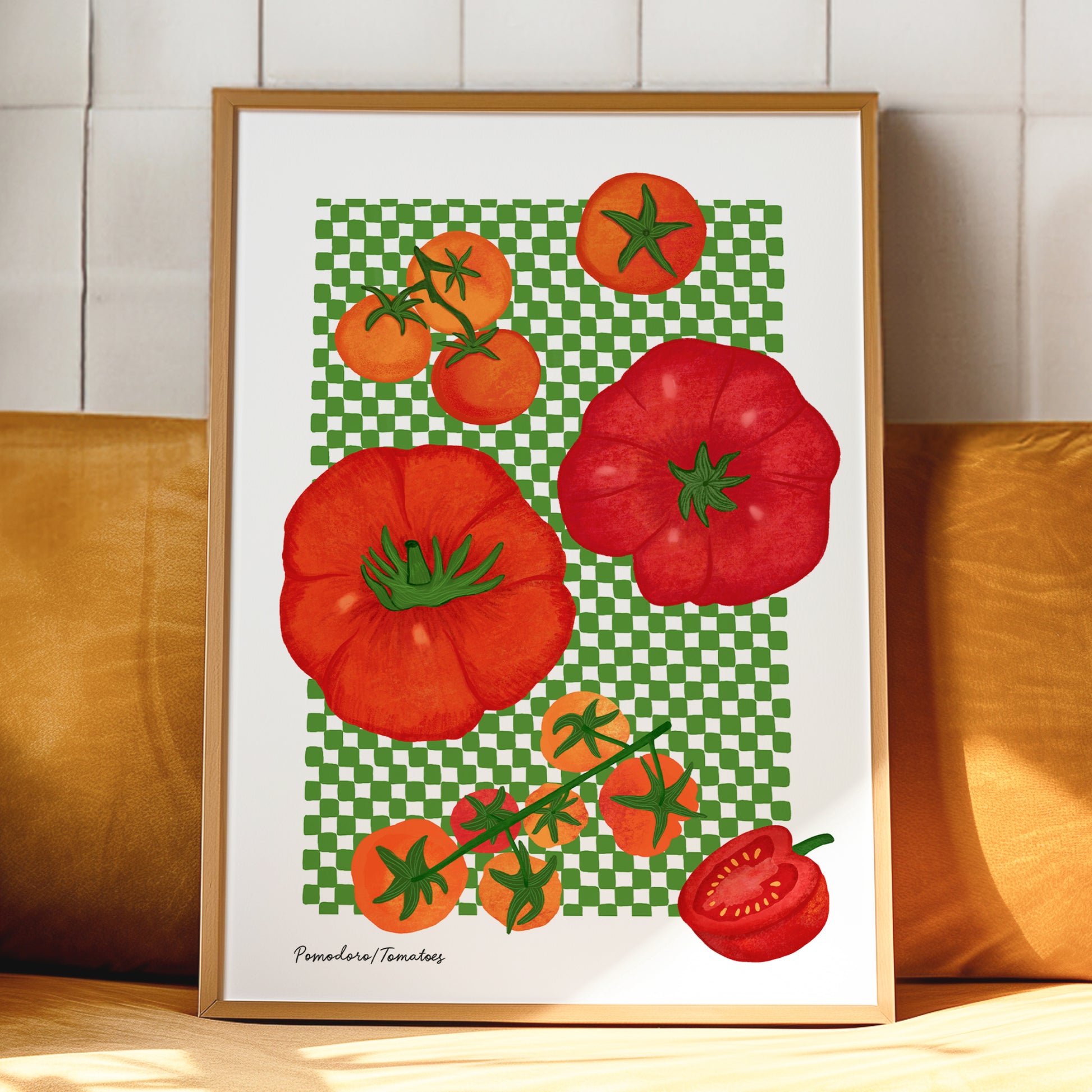 A framed art print featuring a colorful illustration of red and orange tomatoes on a green checkered background, leaning against a yellow cushion in front of a white tiled wall.